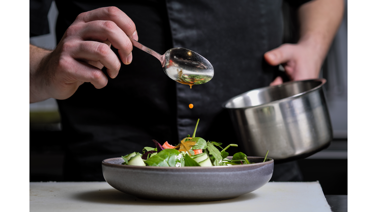 Close-up of the hands of a male chef on a black background. Pour sauce from the spoon on the salad dish.