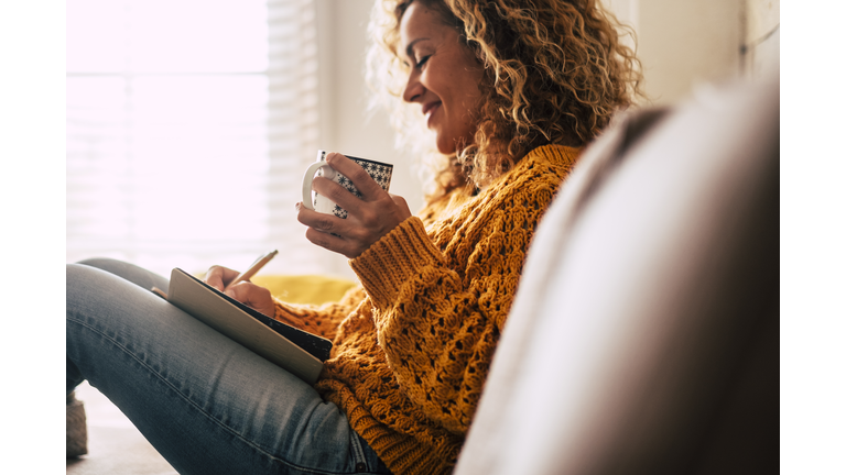 Happy cute lady at home write notes on a diary while drink a cup of tea and rest and relax taking a break. autumn colors and people enjoying home lifestyle writing messages or lists. Blonde curly beautiful lady sit down in the house