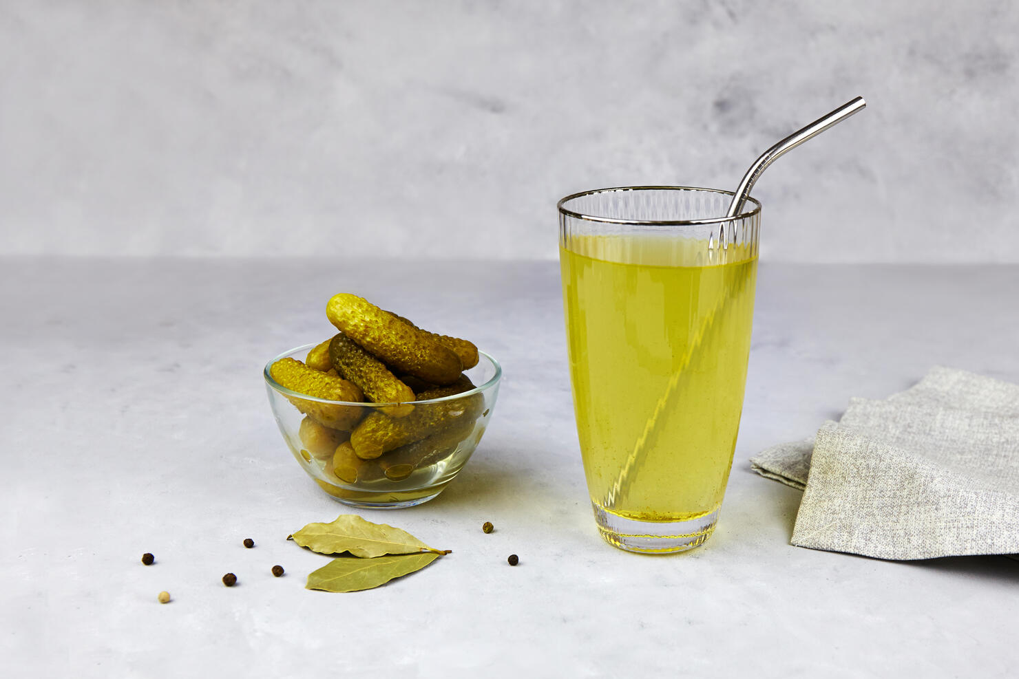 Cucumber pickle or pickle juice in glass with a metal tube for drinks , a bowl with pickled gherkins on light background. Trend drink, sports nutrition, healthy supplements.