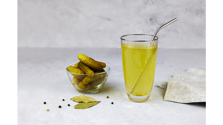 Cucumber pickle or pickle juice in glass with a metal tube for drinks , a bowl with pickled gherkins on light background. Trend drink, sports nutrition, healthy supplements.