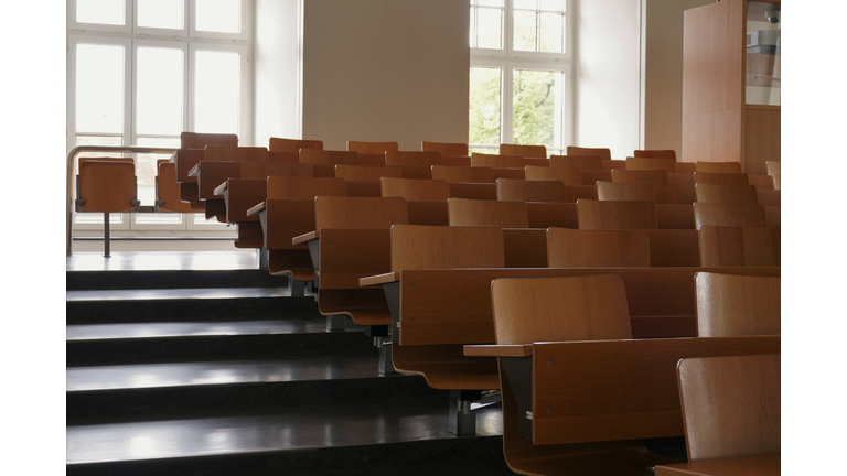 Empty auditorium at university with wooden chairs and banks and large windows and stairs on one side.