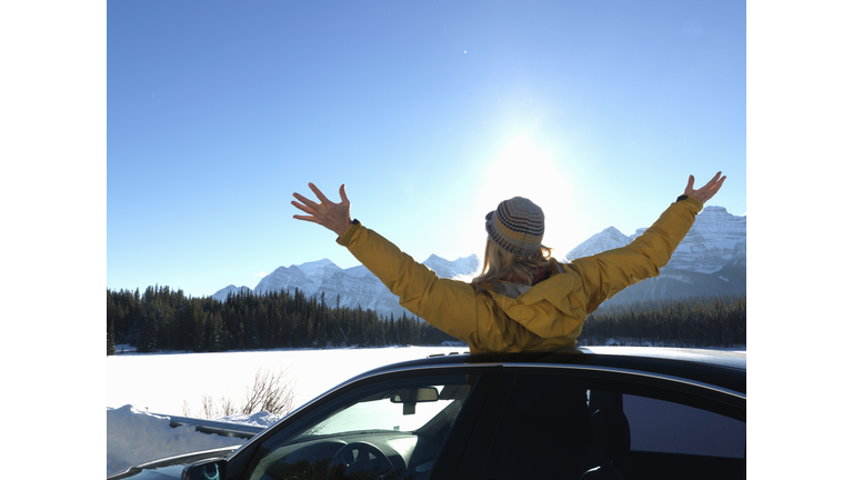 Woman standing in car's sunroof with outstretched arms, rear view