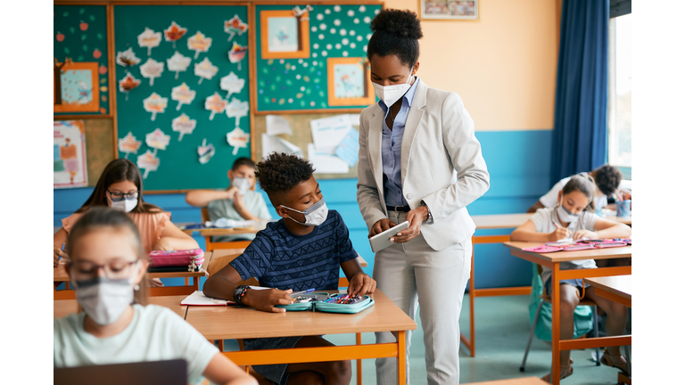 African American teacher and her student using touchpad and wearing protective face masks due to coronavirus pandemic.