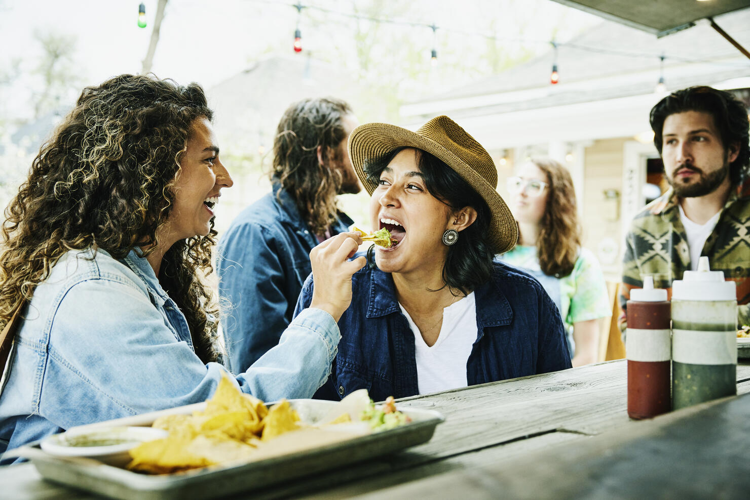 Medium shot of laughing woman feeding friend chip with guacamole while eating at food truck