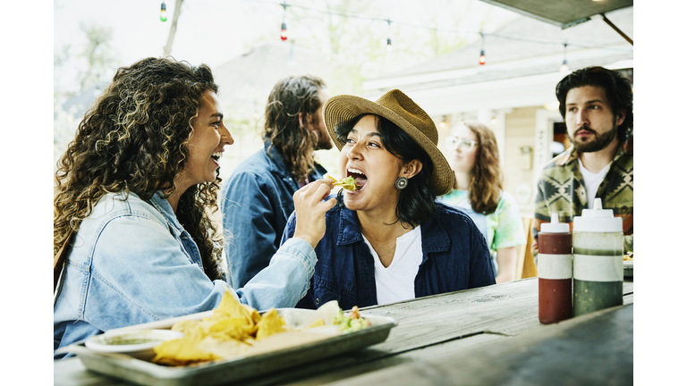 Medium shot of laughing woman feeding friend chip with guacamole while eating at food truck