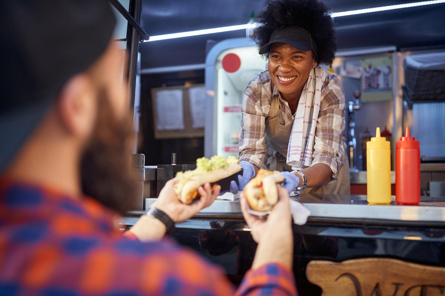 polite afro-american female employee holding, giving, two sandwiches to a beardy caucasian customer. fast food service