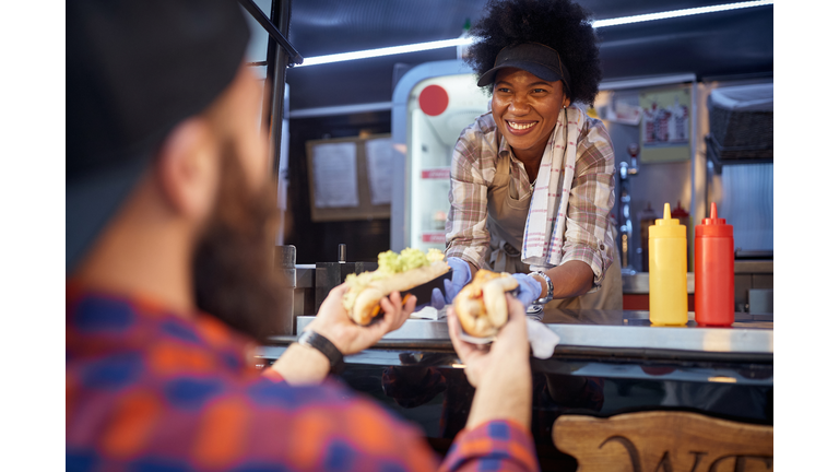 polite afro-american female employee holding, giving, two sandwiches to a beardy caucasian customer. fast food service