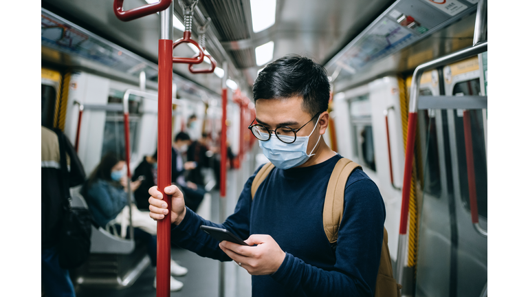 Young Asian man with protective face mask using smartphone while commuting in the city riding on subway