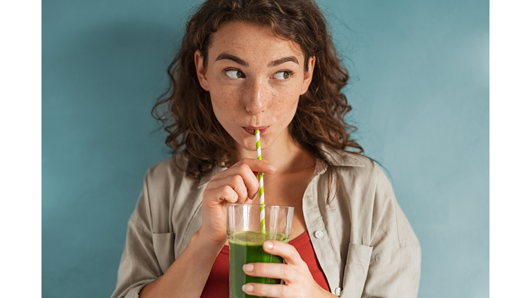 Young woman drinking detox juice with straw on blue wall