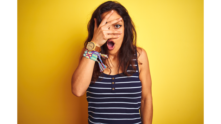 Young beautiful woman wearing striped t-shirt standing over isolated yellow background peeking in shock covering face and eyes with hand, looking through fingers with embarrassed expression.
