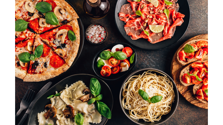 Full table of italian meals on plates Pizza, pasta, ravioli, carpaccio. caprese salad and tomato bruschetta on black background.