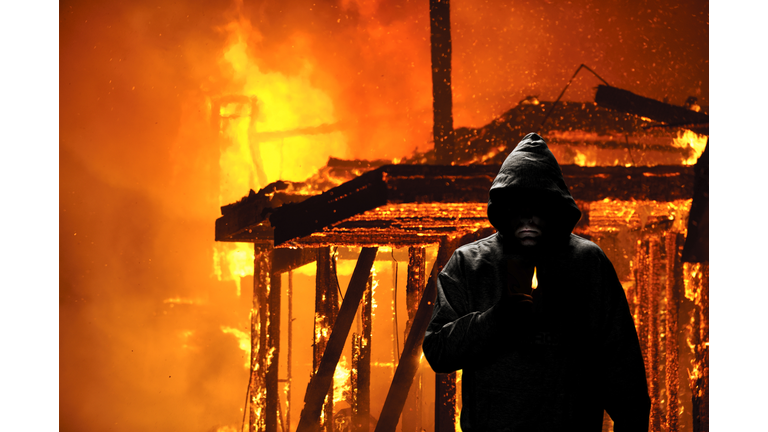 Hooded person holding a lighter in front of burning house