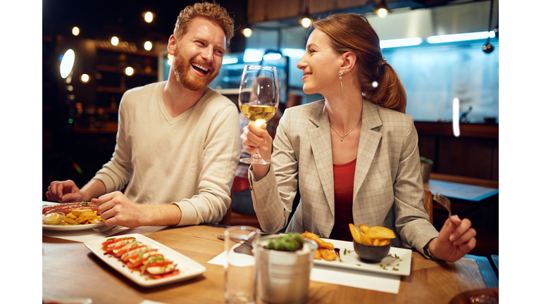 Smiling cheerful couple sitting in a restaurant, having dinner and chatting. Man talking to a woman while a woman listening to him and drinking white wine.