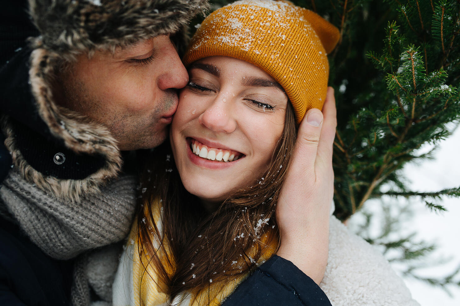 Man kissing his woman in a cheek in front of a fir tree in the winter.