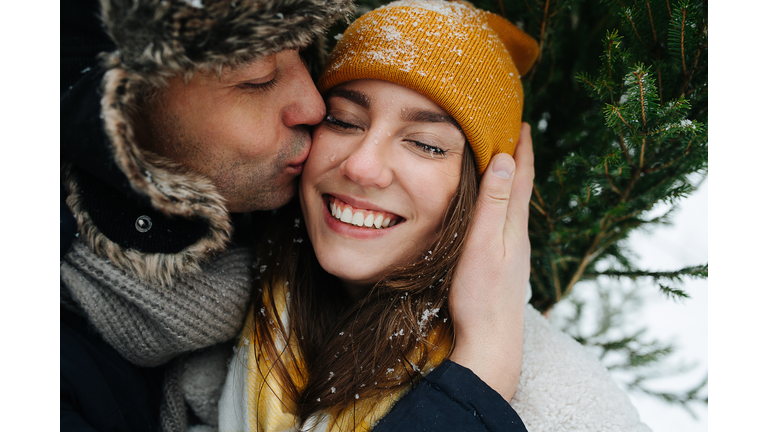 Man kissing his woman in a cheek in front of a fir tree in the winter.