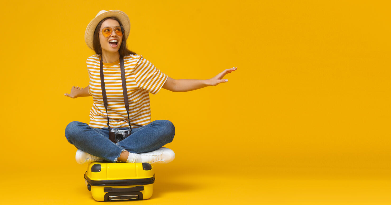 Dreams about traveling concept. Horizontal banner of young tourist girl sitting on suitcase, pretending flying on a plane, isolated on yellow background with copy space.