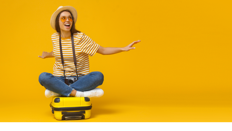 Dreams about traveling concept. Horizontal banner of young tourist girl sitting on suitcase, pretending flying on a plane, isolated on yellow background with copy space.