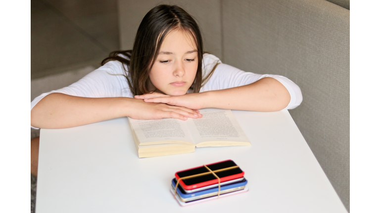 Social media and gadget detox. Sad preteen girl reading book and looking at pile of smartphones at table taken from her. No phone punishment.