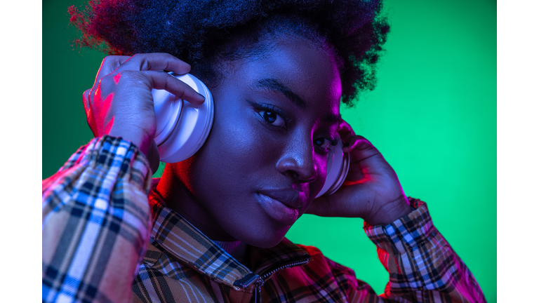 Close-up happy young girl, student listening to music in headphones isolated on dark green studio background in purple neon light.