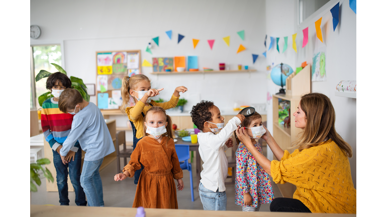 Pre school teacher helping little girl to wear face mask indoors at nursery school, coronavirus concept.