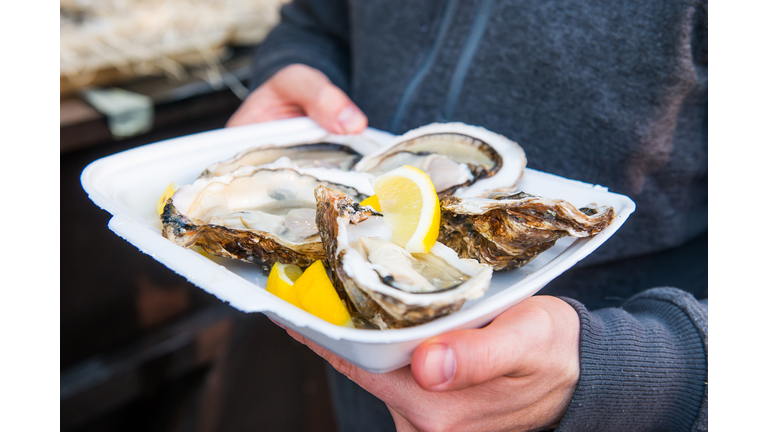 Close up male hand holding take away food tray with fresh opened oyster and lemon slices at street food market, festival, event. Selective focus. copy space.