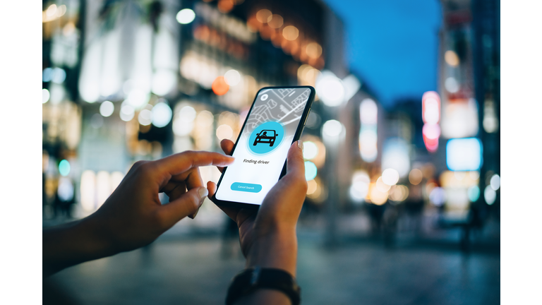 Close up of young woman using mobile app device on smartphone to arrange a taxi ride in downtown city street, with illuminated city traffic scene as background