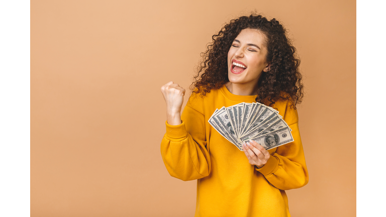 Portrait of a cheerful young woman holding money banknotes and celebrating isolated over beige background.