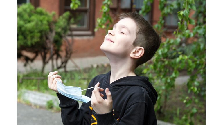 Happy and relaxed boy takes off the protective mask and smiles with closed eyes. Outdoors, spring sunny day.