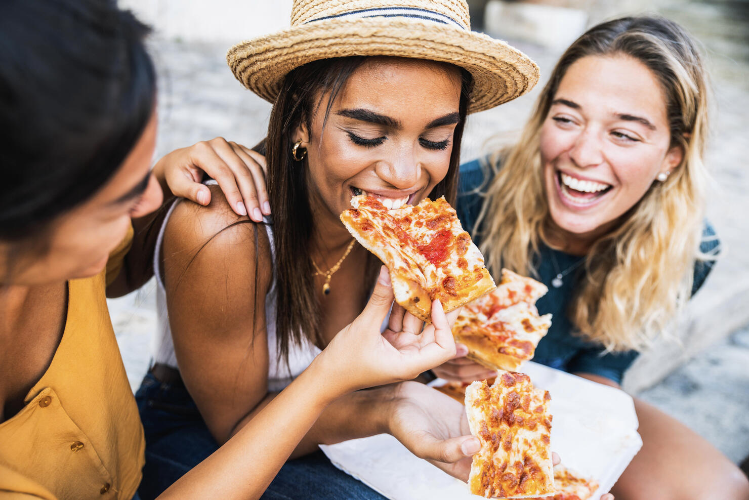 Three young female friends sitting outdoor and eating pizza - Happy women having fun enjoying a day out on city street - Happy lifestyle concept