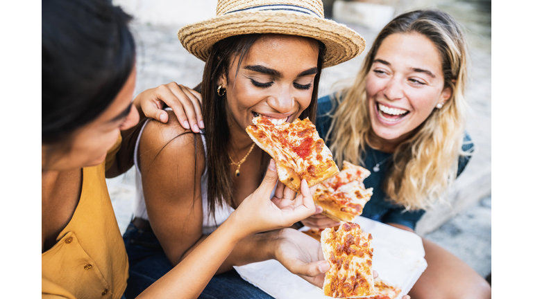 Three young female friends sitting outdoor and eating pizza - Happy women having fun enjoying a day out on city street - Happy lifestyle concept