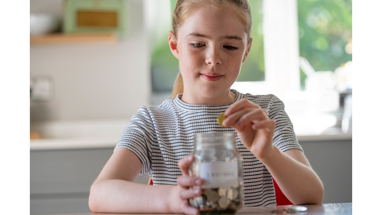 Girl Putting Coins Into Glass Jar Labelled Savings At Home