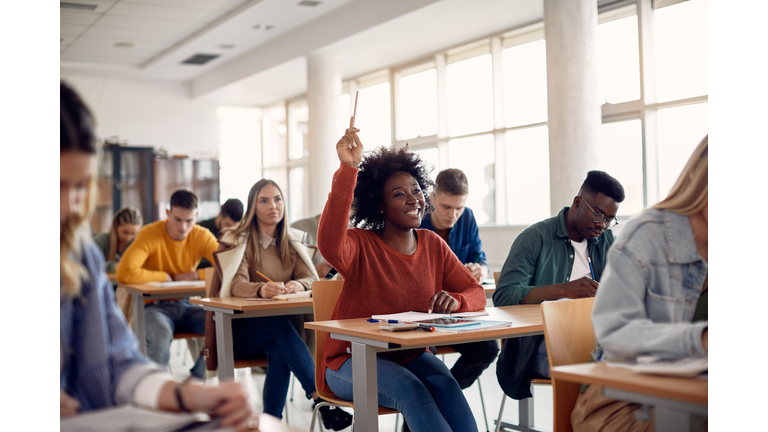 Happy black student raising arm to answer question while attending class with her university colleagues.