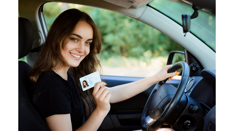 Smiling young female with pleasant appearance shows proudly her drivers license, sits in new car, being young inexperienced driver, looks with joyful expression