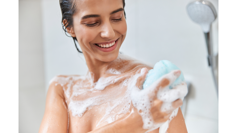 Cheerful young woman using exfoliating loofah while taking shower