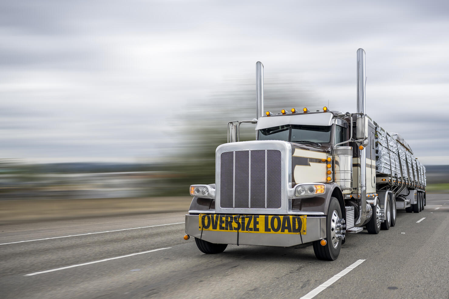 Powerful classic dark big rig semi truck with oversize load sign on the front transporting lumber on the flat bed semi trailer running on the wide highway road