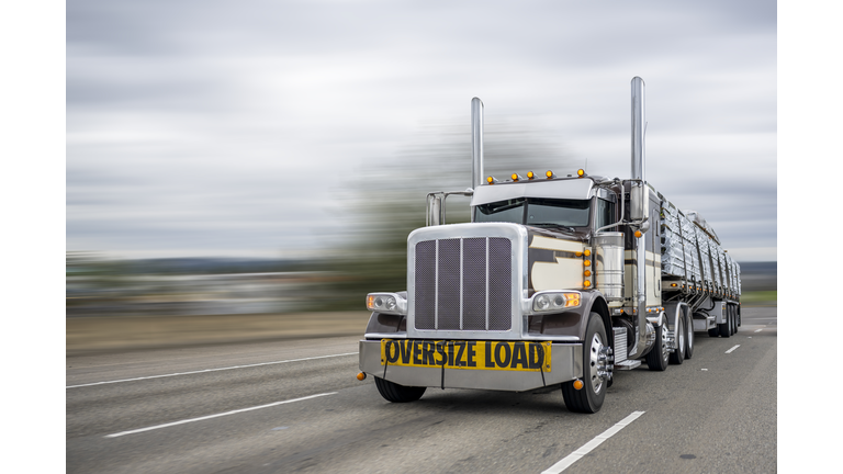 Powerful classic dark big rig semi truck with oversize load sign on the front transporting lumber on the flat bed semi trailer running on the wide highway road