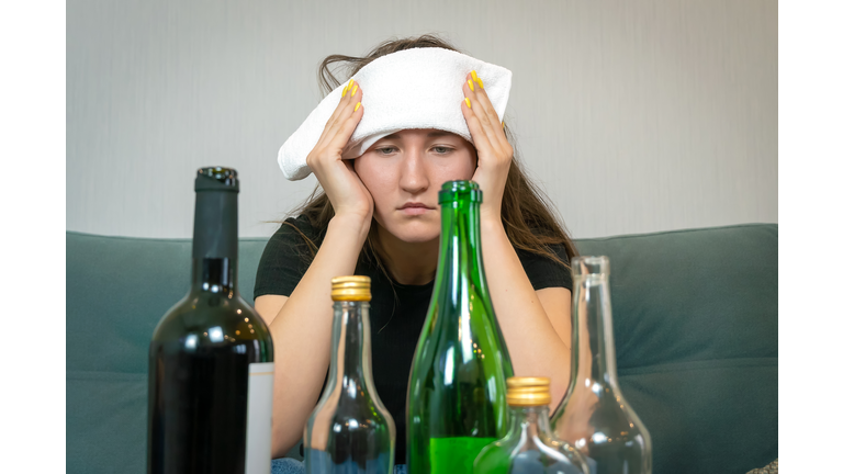 Young woman with a white towel on her forehead from a headache sits in front of empty bottles of alcohol. Concept of Monday morning, morning after drinking alcohol