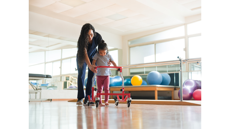 Physical therapist helping girl to walk with wheeled frame