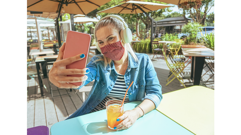 Girl in bar takeing a selfie with a smartphone with her face mask on as a protection for coronavirus time - Teenager chilling outside and enjoying a cold drink - Lifestyle of covid-19