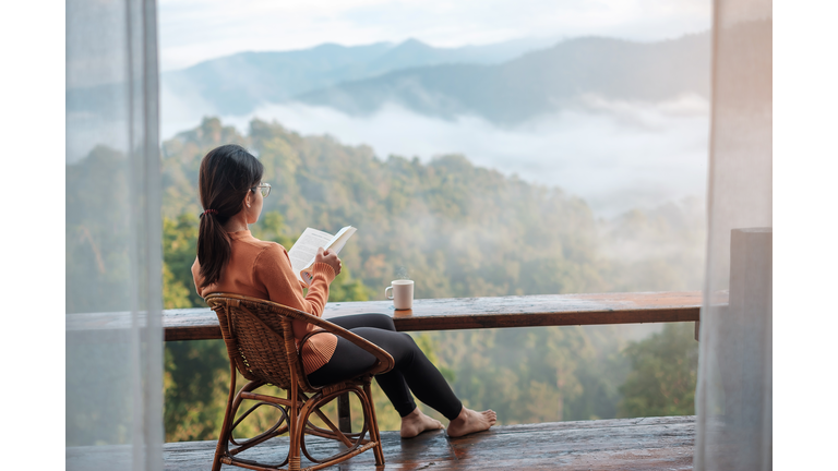 young woman reading book near window and looking mountain view at countryside homestay in the morning sunrise. SoloTravel, journey, trip and relaxing concept