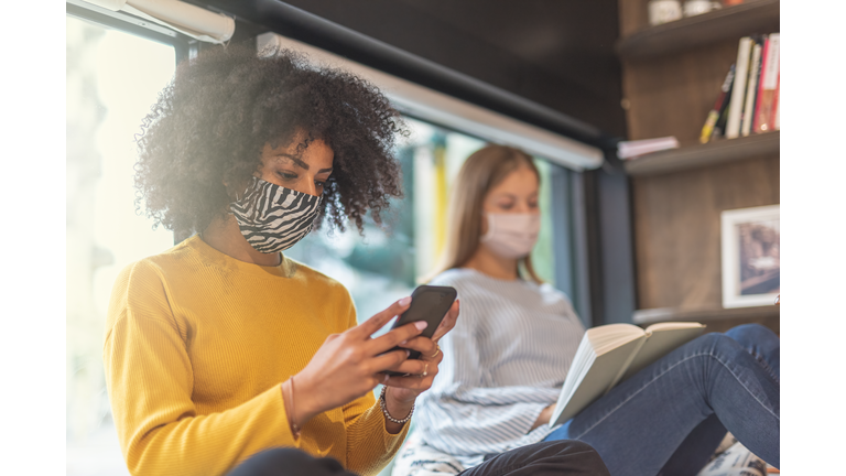 a young African-American woman looks on her smart phone at the latest news of coronavirus infection, two students in a bookstore with face masks to prevent the contagion of covid19, sun flare effect