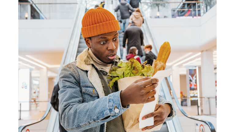 African man with a bag of groceries looks surprised and upset at a receipt from a supermarket with high prices. The rise in the price of food