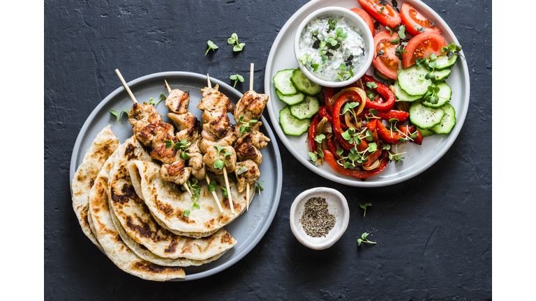 Mediterranean style lunch table - turkey skewers, flatbread, tomatoes, cucumber salad, baked sweet pepper, yogurt herb sauce  on a dark background, top view