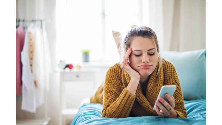 Bored young woman with smartphone lying on bed indoors at home.