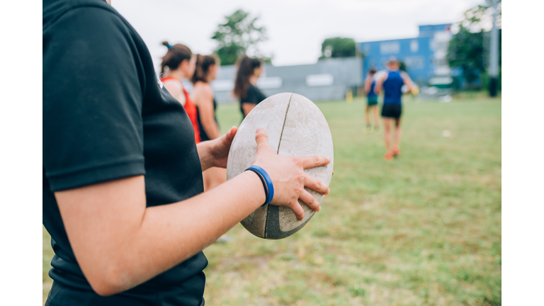A group of women and their male coach at rugby training, one woman holding a rugby ball.