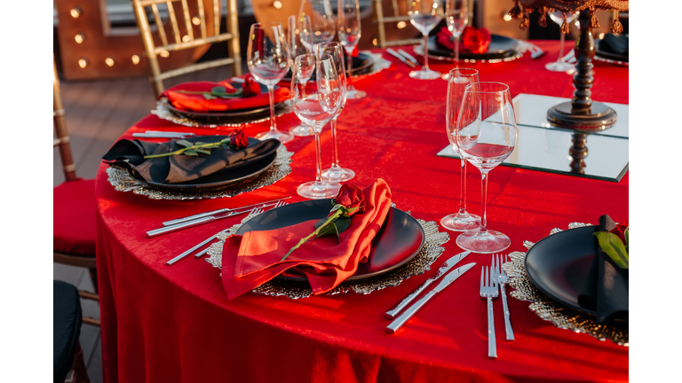 Guest table setting for banquet in black, red and gold style. Elegant and luxury dinner arrangement: decor, tablecloth, plates with napkins and fresh roses, glasses, cutlery. Themed party celebration