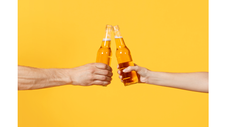 Close up cropped of woman and man two hands horizontal holding lager beer glass bottles and clinking isolated on yellow background. Sport fans cheer up. Friends leisure lifestyle concept. Copy space.