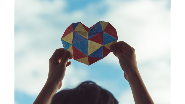 Child hands holding colorful paper heart over blue sky background. World autism awareness day concept