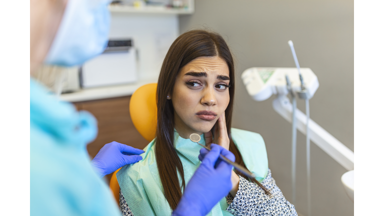 Young woman holding cheek in chair at dentist, having toothache. Shot of a young woman suffering from toothache while sitting in the dentist"u2019s chair