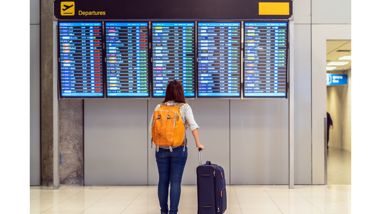 Back side of traveler with luggage standing over the flight board for check-in at the flight information screen in modern an airport, travel and transportation with technology concept.